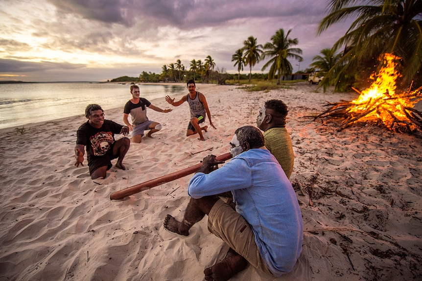 Five men with face paint and didgeridoo sitting on a beach in front of a bonfire