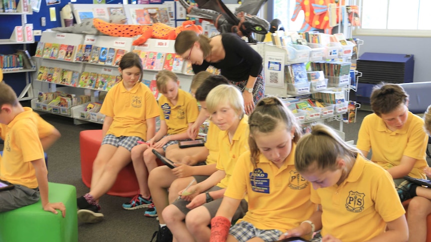 Children sit in their school library working on their iPads. Teacher is in the photo