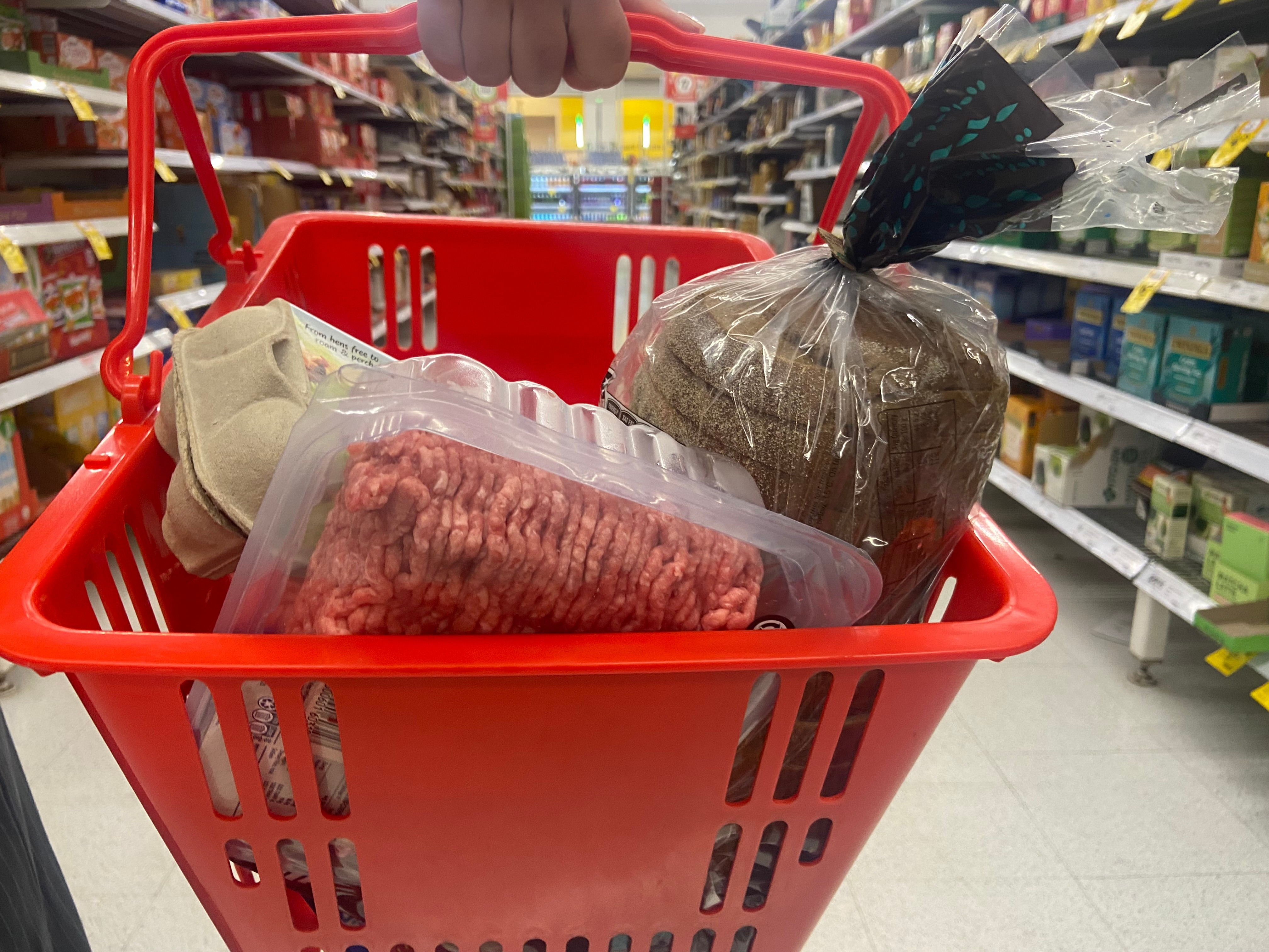 A red basket containing eggs, bread and mince, in a shopping aisle