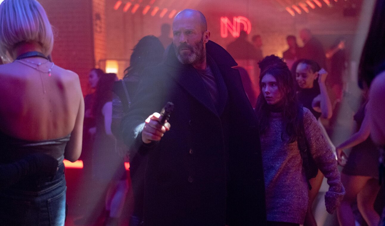 Jason Statham in a tense standoff, shielding a young girl amid shadowy urban threats in the thriller film Shelter