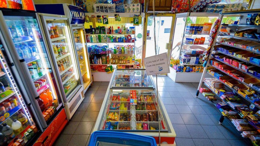 The interior of a milk bar, with drink and ice cream refrigerators and shelves stacked with chocolates and other groceries.