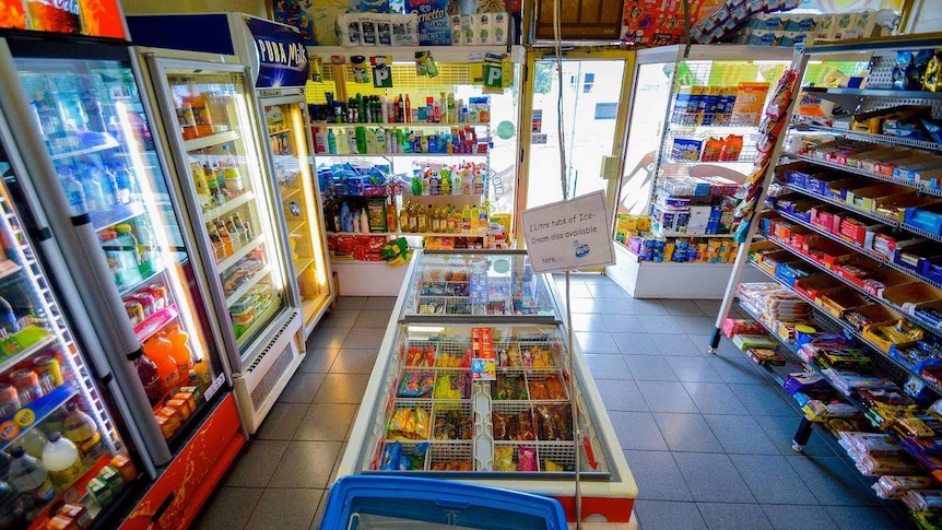 The interior of a milk bar, with drink and ice cream refrigerators and shelves stacked with chocolates and other groceries.