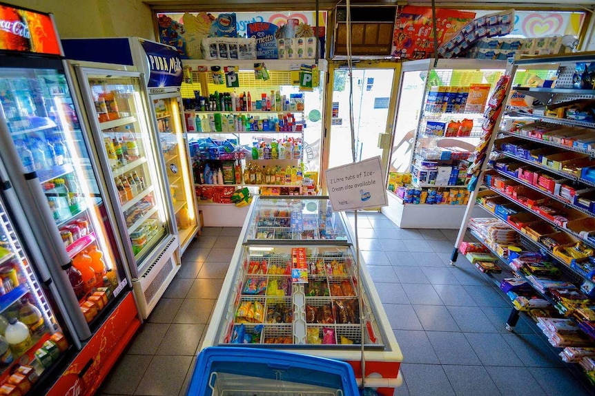 The interior of a milk bar, with refrigerators for drinks and ice cream, and shelves stacked with chocolate and other groceries.