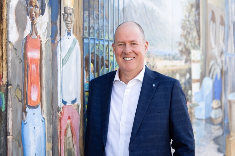 Smiling, shaved-headed man wearing shirt and blazer and standing in front of a mural