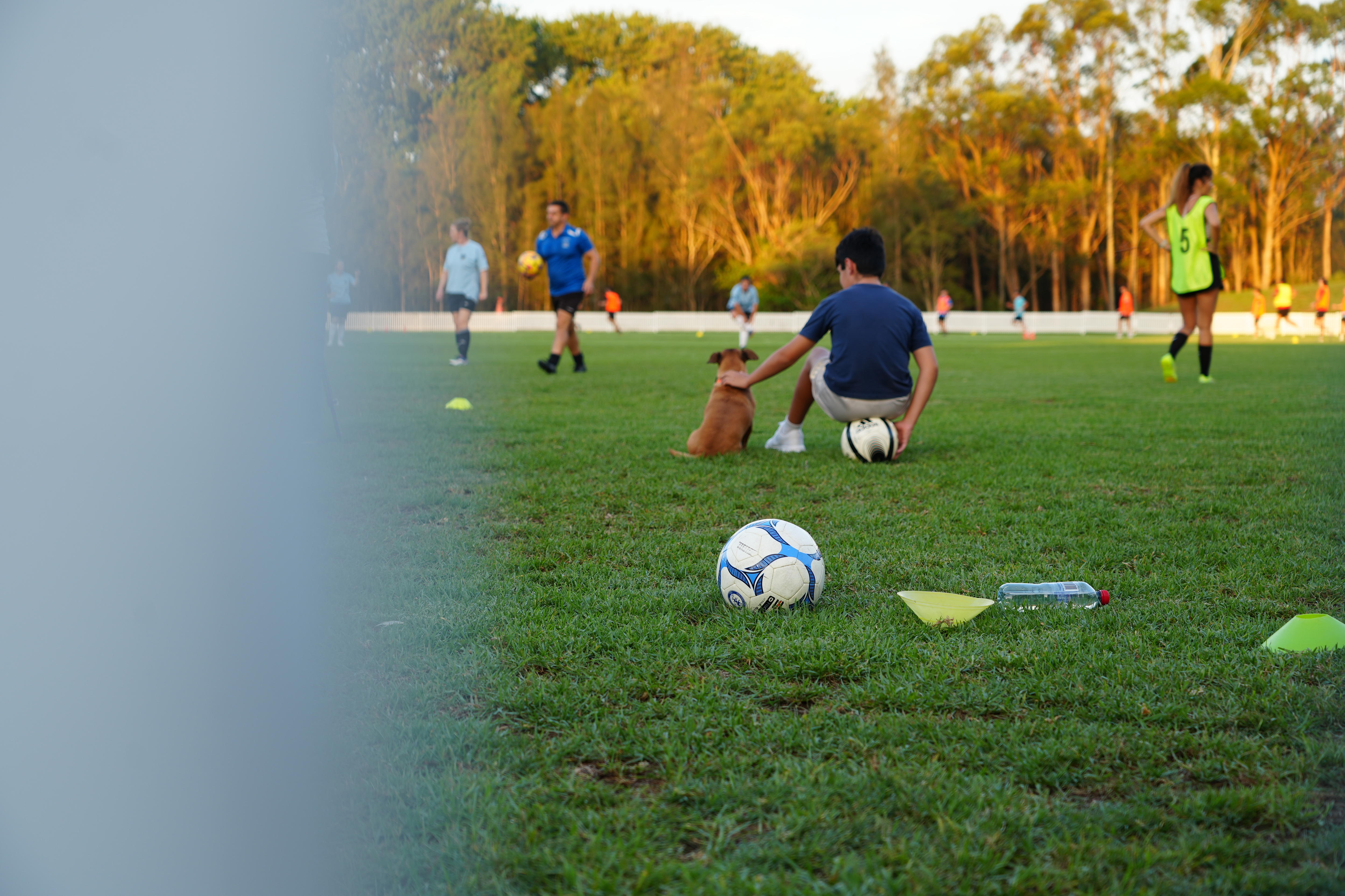 Um menino senta em uma bola de futebol e dá tapinhas em um cachorro, enquanto observa mulheres treinando. 