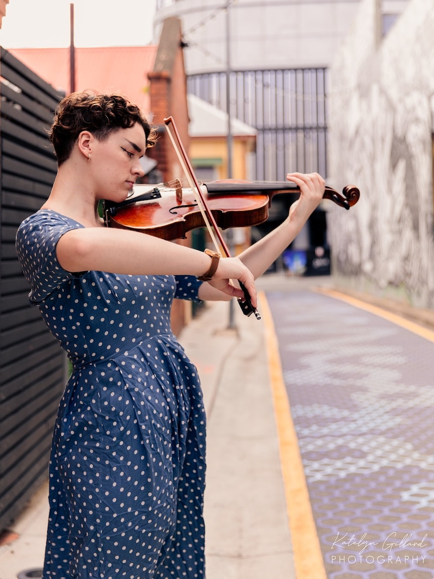 An side on image of Sophia Mackson in a blue polka dot dress in urban lane way playing viola.