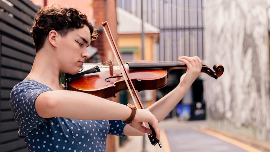 An side on image of Sophia Mackson in a blue polka dot dress in urban lane way playing viola 