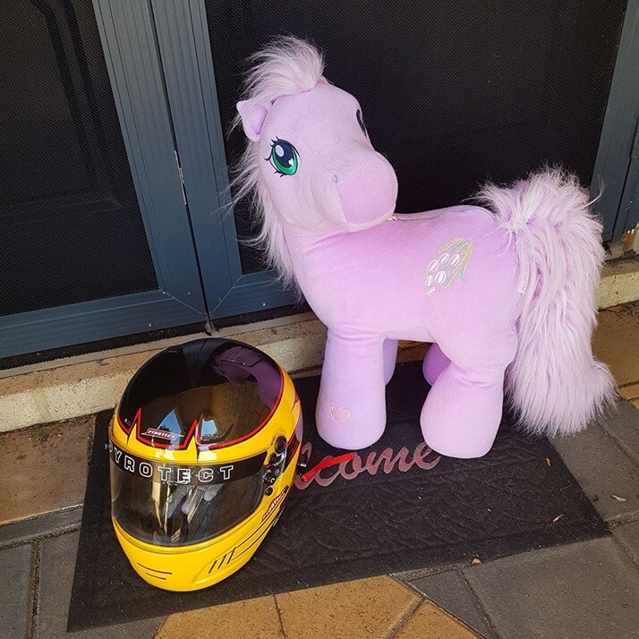 A yellow and black motor racing helmet sits on a doormat with a pink pony soft toy next to it.