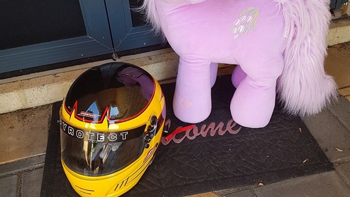 A yellow and black motor racing helmet sits on a doormat with a pink pony soft toy next to it.
