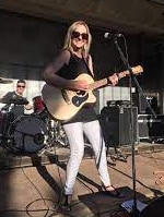 Donna dai capelli biondi in piedi al microfono sul palco con la chitarra e la band dietro di lei