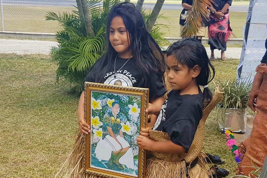 Two Tongan girls in grass skirts hold photo of a man, Tonga airport