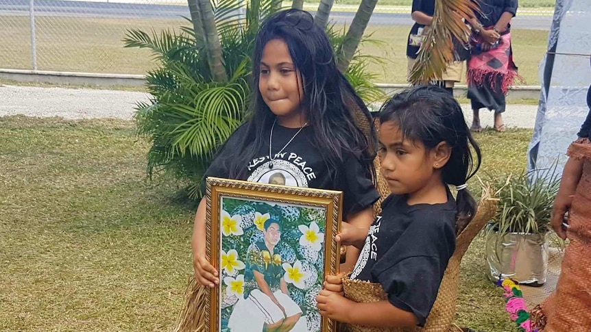 Two Tongan girls in grass skirts hold photo of a man, Tonga airport