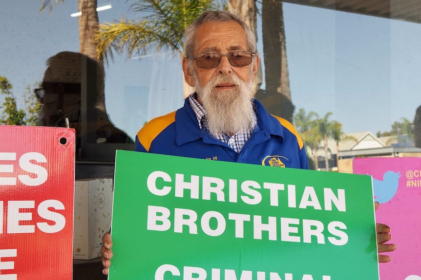 Man with beard stands holding sign that says 'Christian Brothers criminal'.
