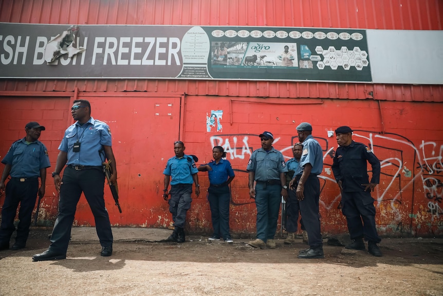 Uniformed PNG police, some armed, stand near the red walll of an industrial building.