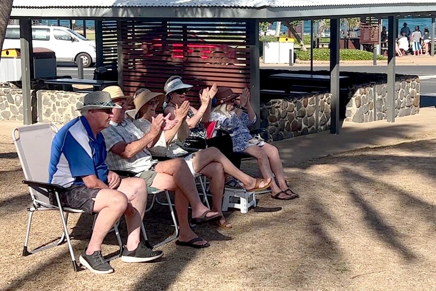 Five people sitting in camping chairs in a line on a grassy area beside a beach, clapping hands