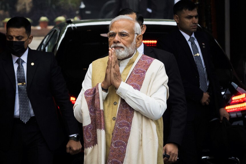 Narendra Modi wearing a white saree holds his hands in prayer while surrounded by security.