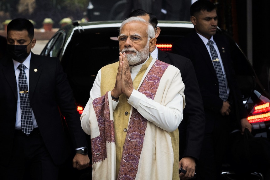 Narendra Modi wearing a white saree holds his hands in prayer while surrounded by security.
