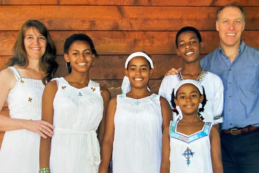Smiling Caucasian man, women with three African girls and one boy. Children and woman wear white, stand against a wooden wall.