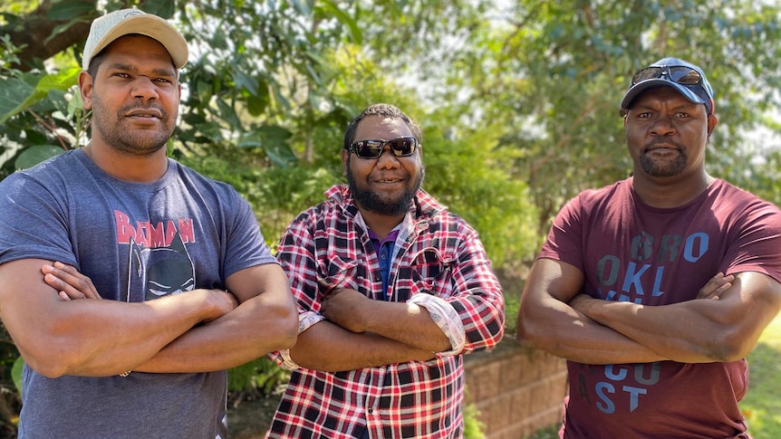 Three Indigenous men stand side by side with arms crossed looking at the camera. They are standing outside in front of a bush. a