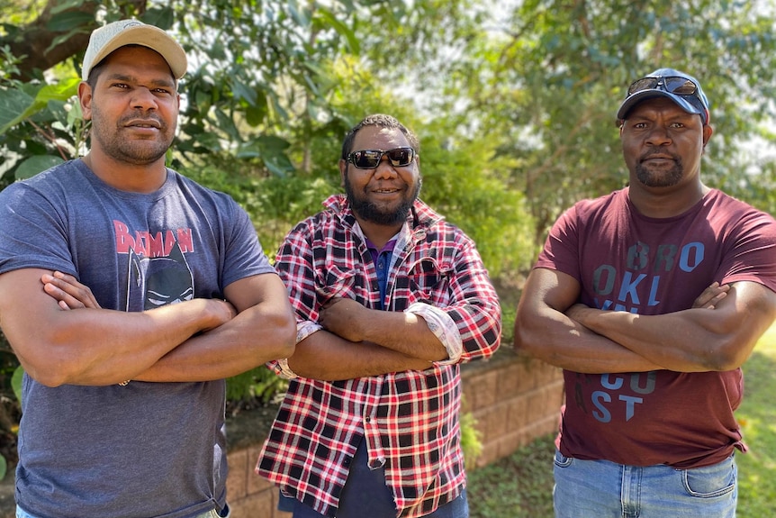 Three Indigenous men stand side by side with arms crossed looking at the camera. They are standing outside in front of a bush. a