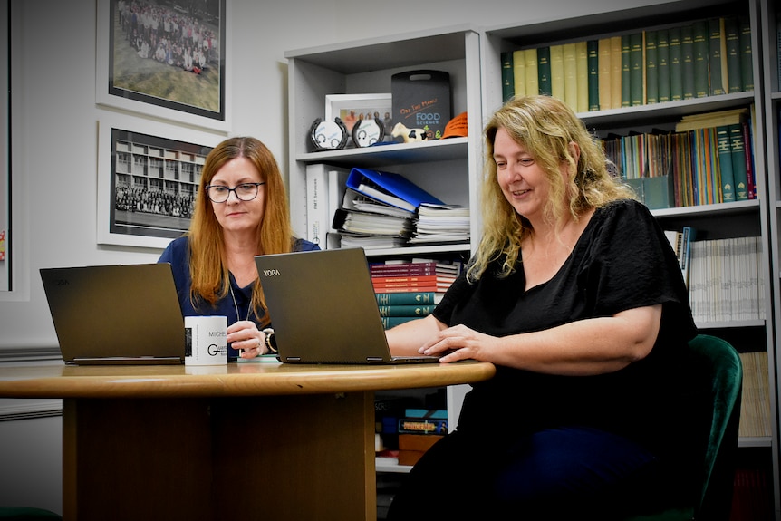 Two women sit in a library in front of two laptops
