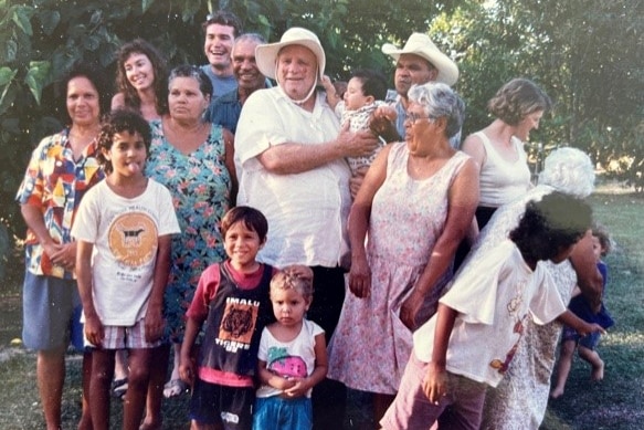 An older man wearing a white shirt and floppy, wide-brimmed hat stands among a large extended family of First Nations people