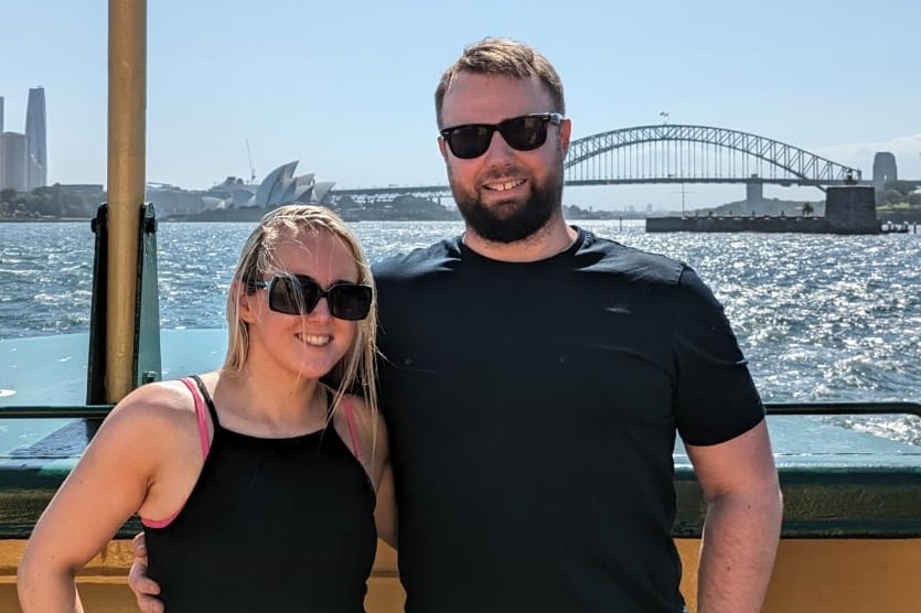 Young blonde woman wearing black top with young man with a beard in front of the Sydney Harbour Bridge.