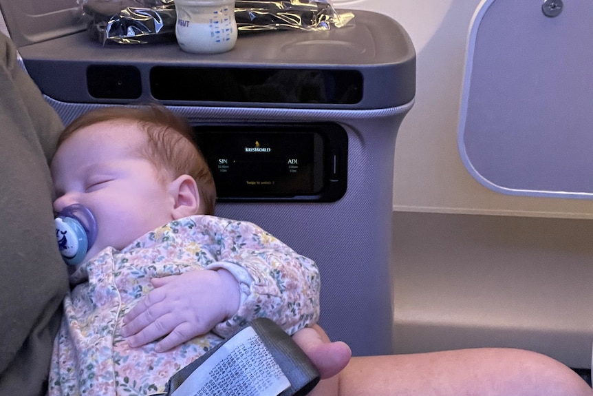 A baby sleeps in her mother's arms on a flight with her bottle on a ledge behind her.