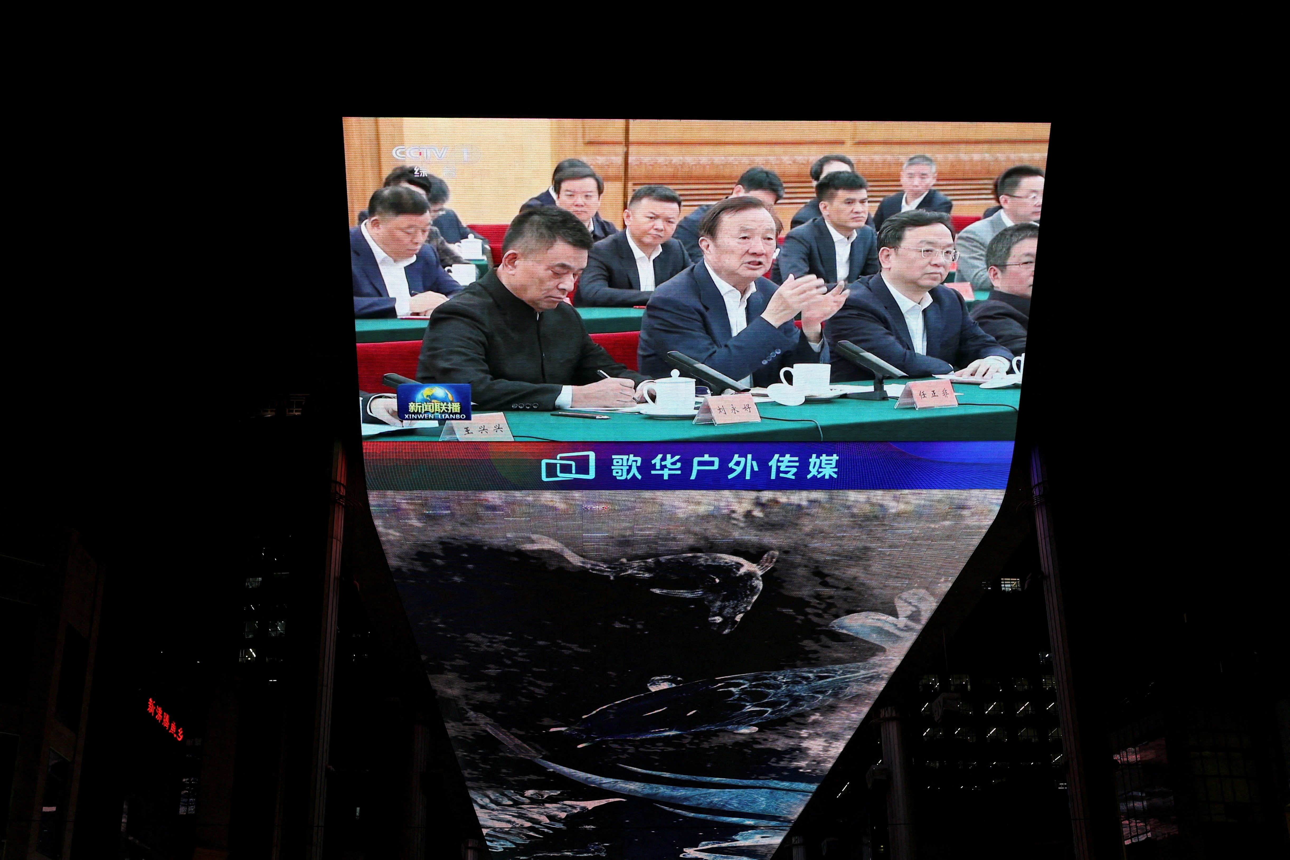 A giant screen projected in a shopping complex showing Chinese businessmen speaking at a formal hearing.
