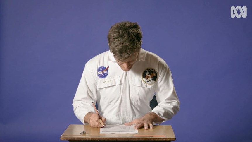 Man in astronaut overalls sits at table, writes on document