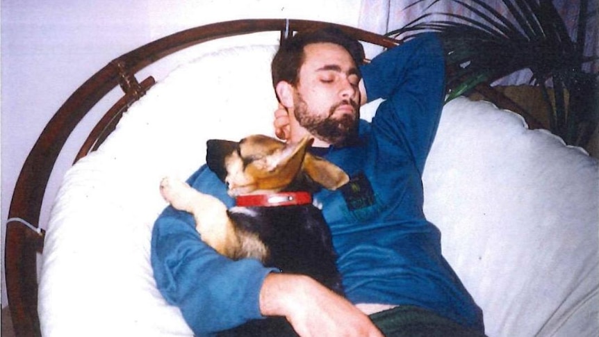 A young Bradley Robert Edwards reclines on a wicker sofa, cuddling a small black and tan dog, with his eyes closed.