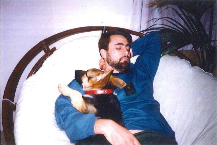 A young Bradley Robert Edwards reclines on a wicker sofa, cuddling a small black and tan dog, with his eyes closed.