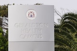 A grey sign with Geraldton Health Campus embossed on it. Below is red text saying hospital. There are palm trees behind it.