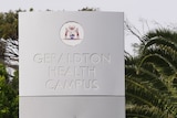 A grey sign with Geraldton Health Campus embossed on it. Below is red text saying hospital. There are palm trees behind it.  