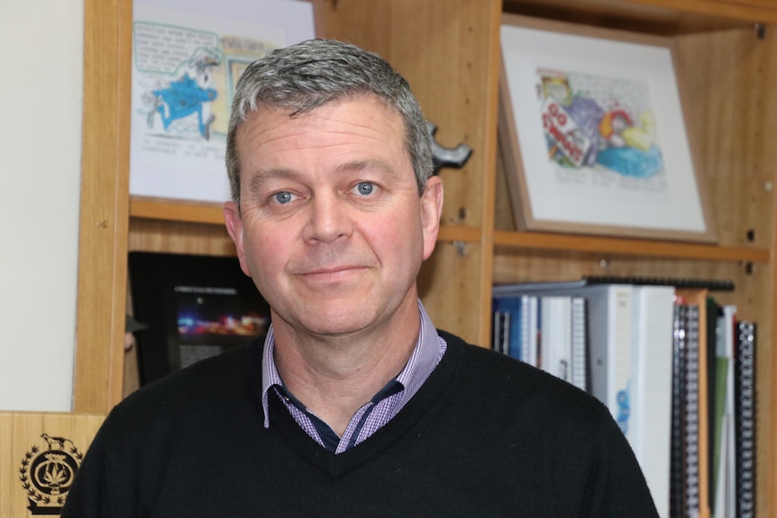 A man standing in front of a bookshelf looks at the camera with a serious expression