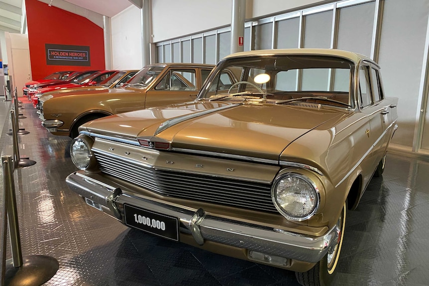 A brown vintage Holden with a '1,000,000' number plate, in the foreground in front of a row of Holdens.