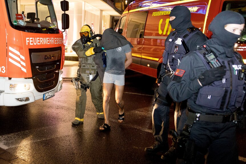 A person with a black hood over their face and hands behind their back is escorted by firemen, next to fire trucks.