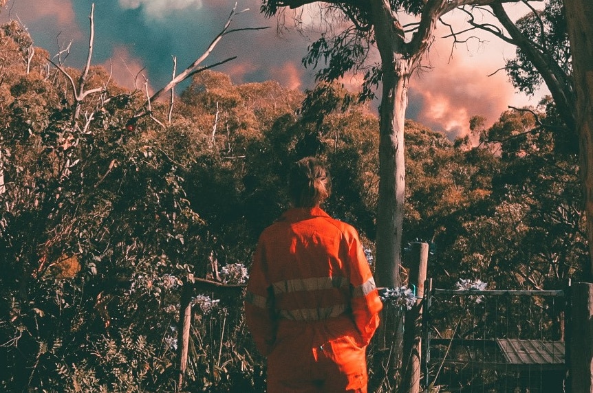 A woman stands wearing red overalls as fire approaches