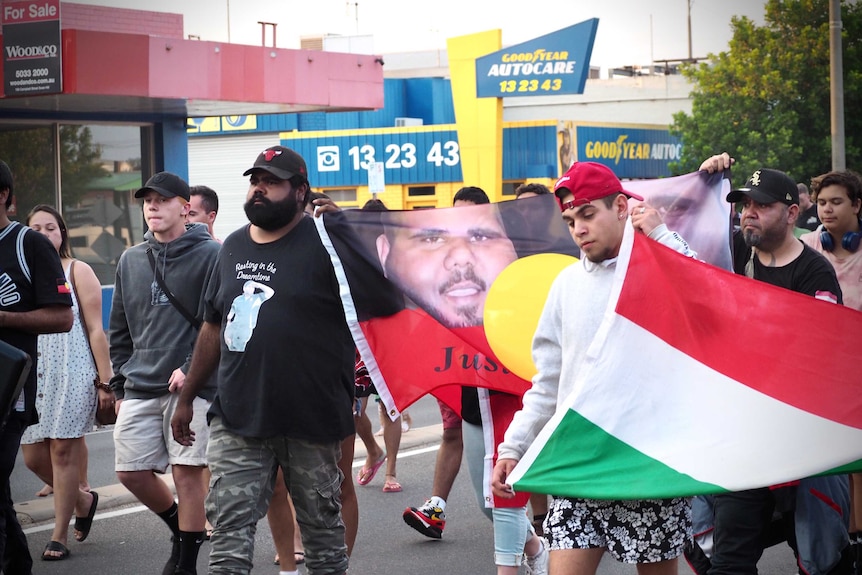 a group of men walking along a street holding a banner bearing the face of the late Jesse Edwards and a Hungarian flag