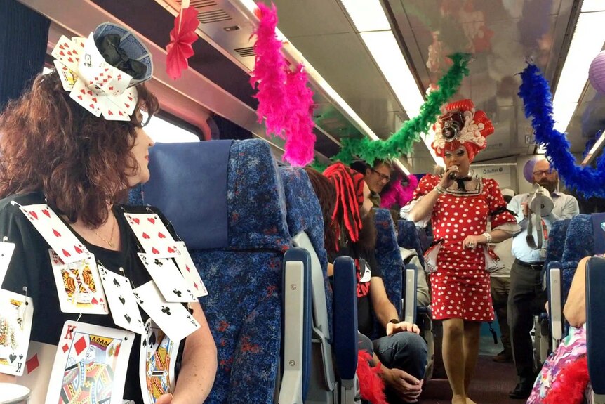 A drag queen dances down the aisle of a train carriage decorated with feather boas.