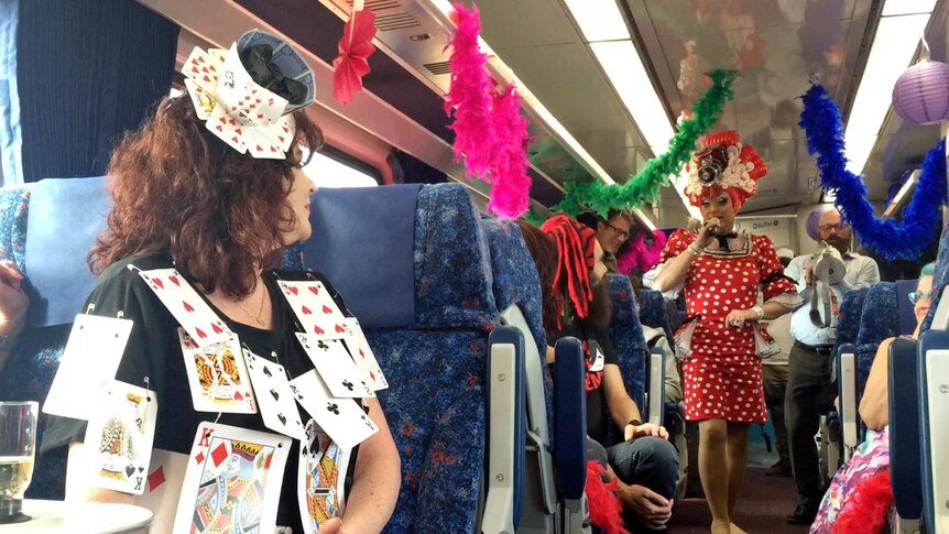 A drag queen dances down the aisle of a train carriage decorated with feather boas.
