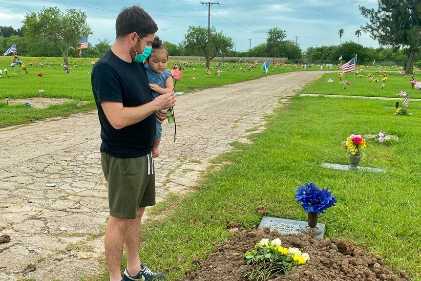 A man in a face mask holding a little girl and a rose stands at the foot of a recently filled gravesite