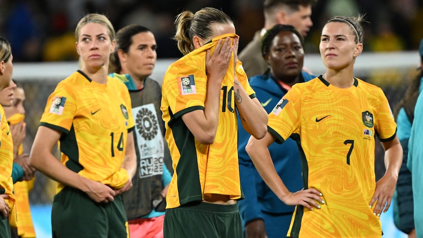A Matildas soccer player rubs her face on her green and gold jersey as other plays look dejected.