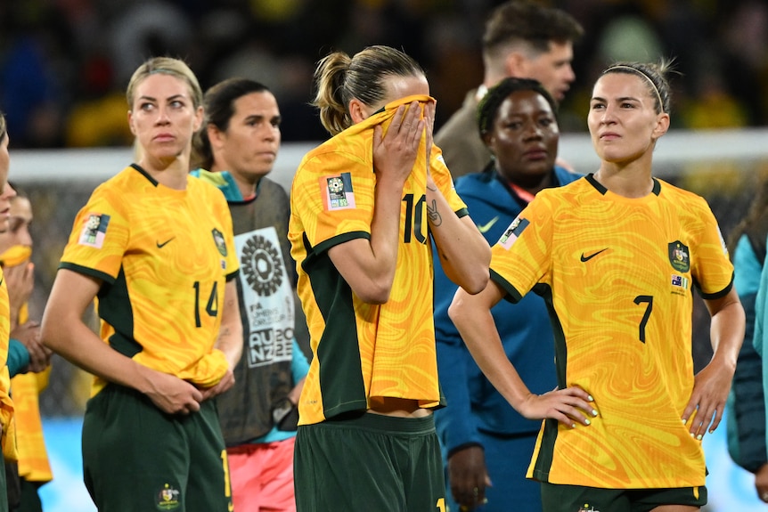 A Matildas soccer player rubs her face on her green and gold jersey as other plays look dejected.