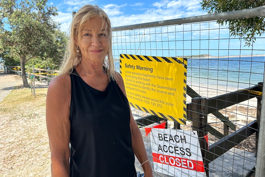 A blonde woman in dark exercise clothing stands next to a warning sign at a beach.