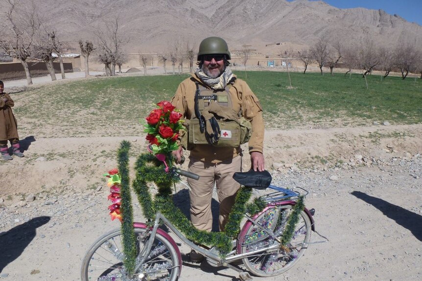 A middle-aged man in combat vest, smiling, with a decorated bicycle.