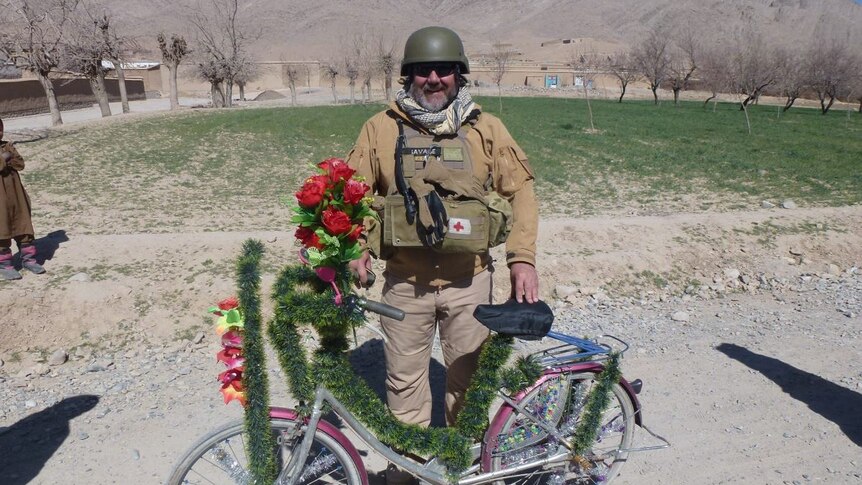 A middle-aged man in combat vest, smiling, with a decorated bicycle.