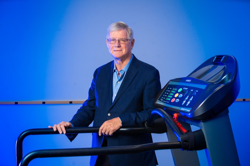 Barry Willer, a man wearing a navy blazer and spectacles, poses for a photo on a black treadmill