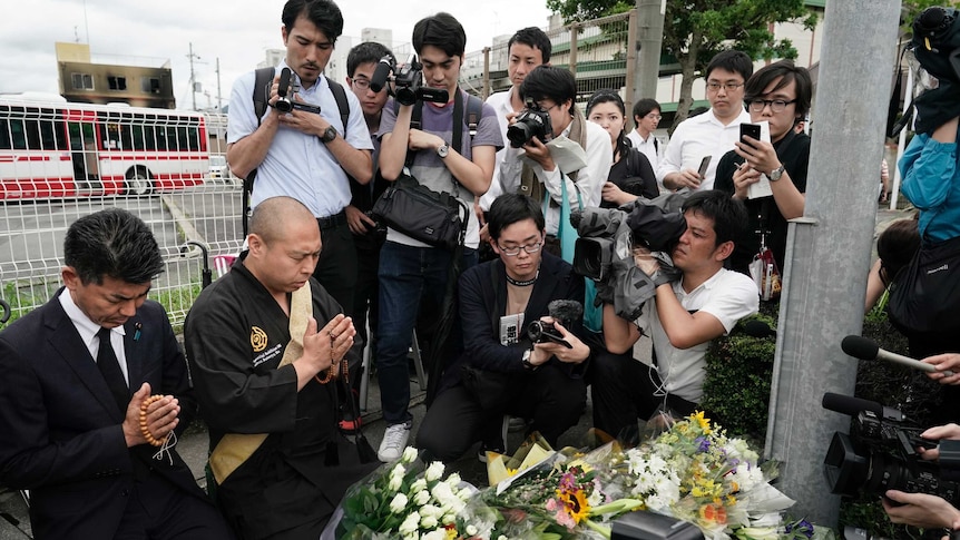 A group of media pray in front of flowers, with a large number of media filming them
