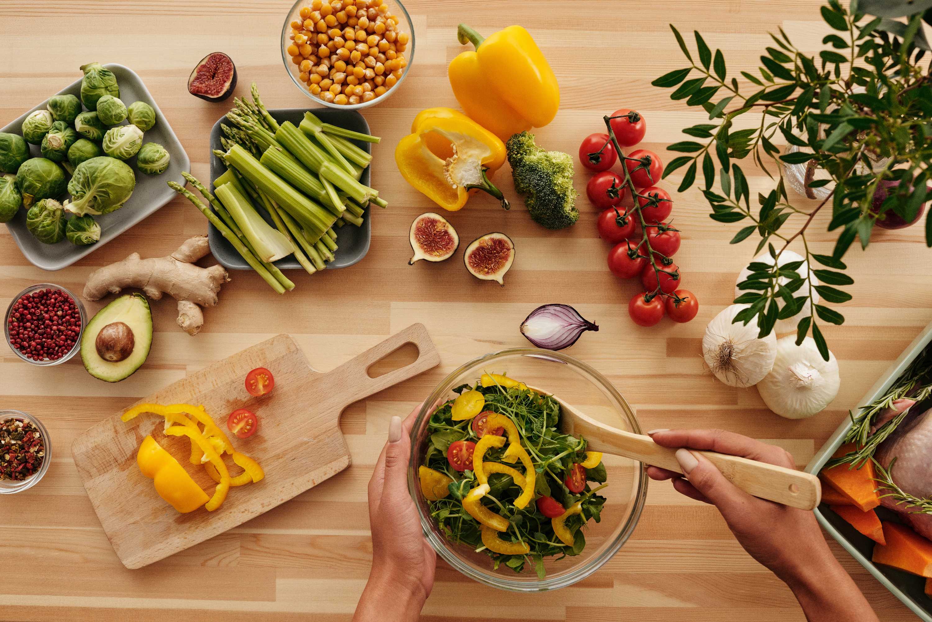 A person stirs a salad on a bench covered with fresh vegetables and legumes.